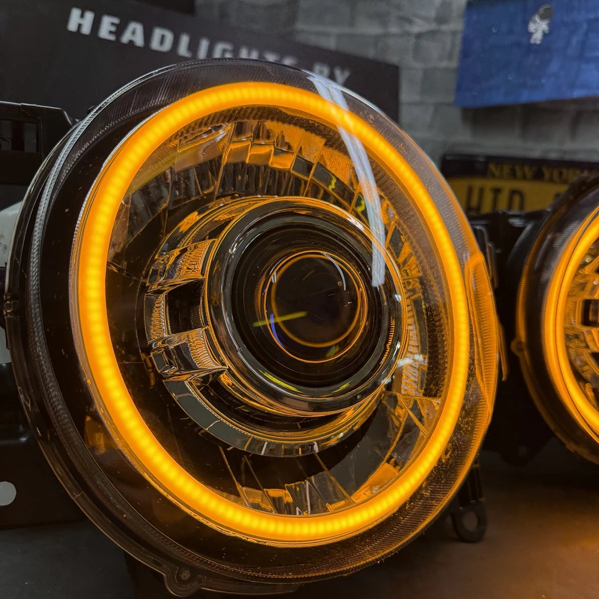 A close-up of a round car headlight with a bright amber/orange LED halo ring illuminated, set against a dark workshop or showroom background with another headlight partially visible on the right.