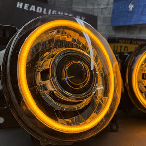 A close-up of a round car headlight with a bright amber/orange LED halo ring illuminated, set against a dark workshop or showroom background with another headlight partially visible on the right.
