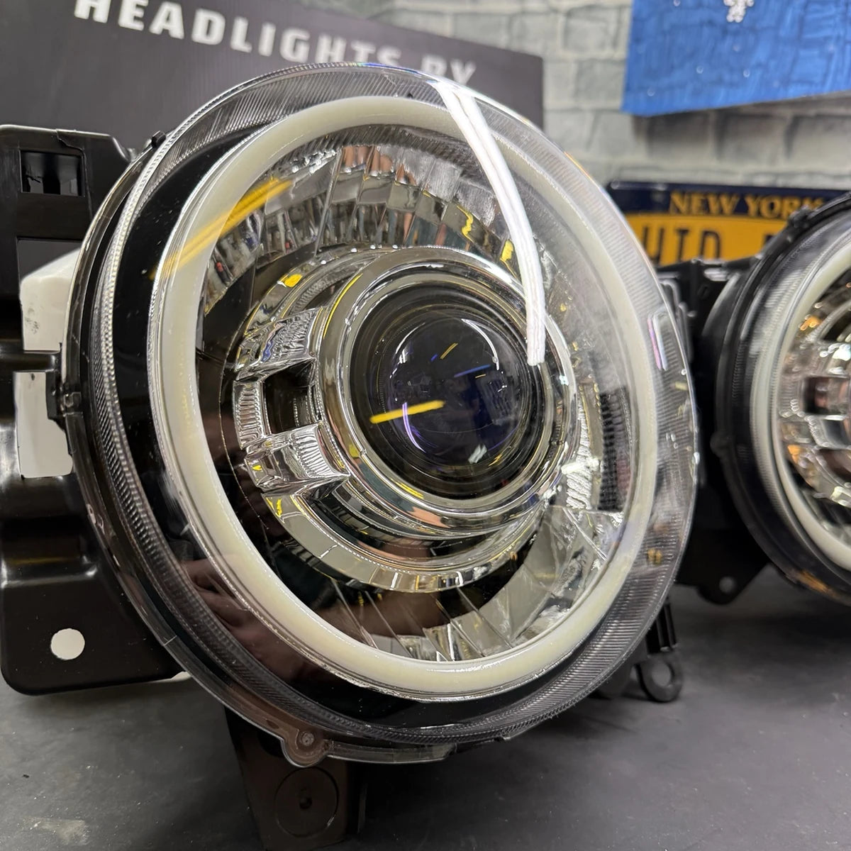 A close-up of a circular automotive headlight assembly with a clear lens, chrome inner ring, and bright white LED ring, photographed in a workshop setting with another headlight partially visible behind it.