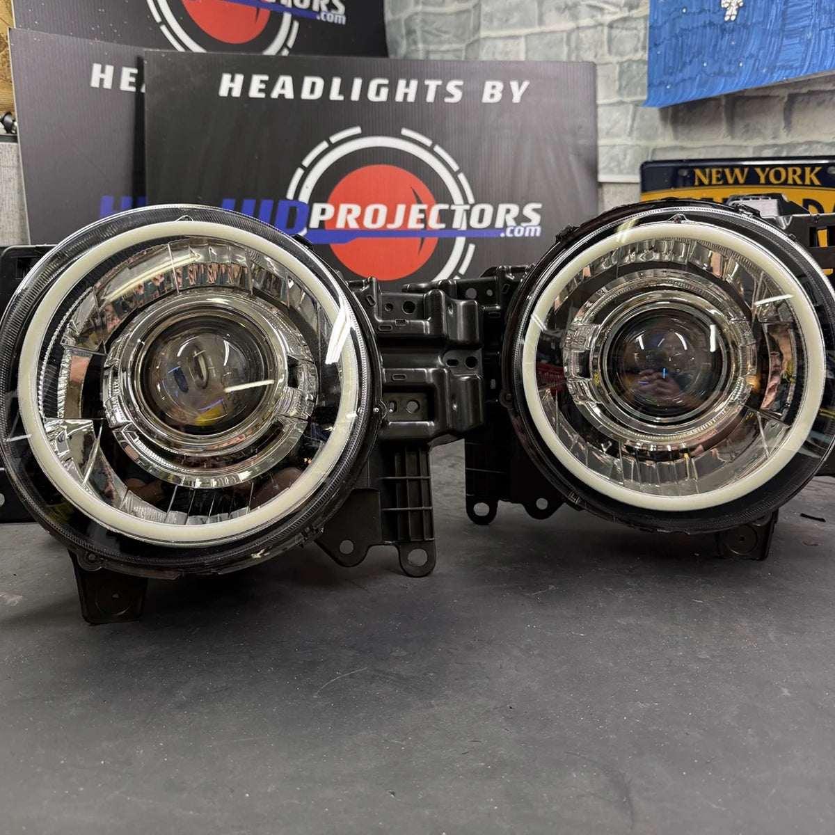 A pair of aftermarket car headlights with clear lenses, white LED rings, and black housings, displayed on a dark surface with a branded background box reading 'HEADLIGHTS BY PROJECTORS'.