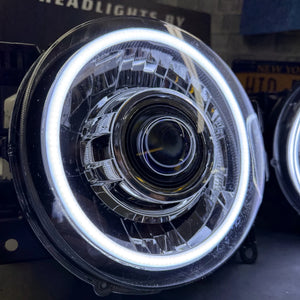 Close-up of a circular car headlight with a bright white LED halo ring and complex multi-element lens assembly, photographed in a workshop setting.