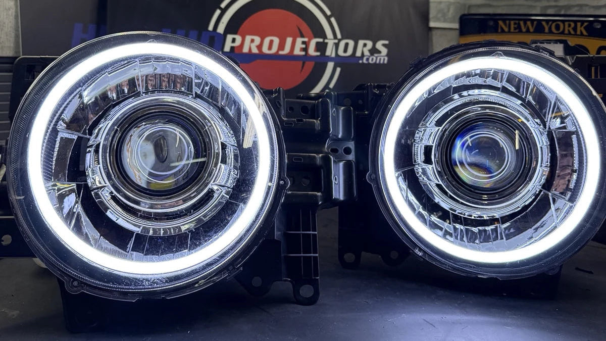 A pair of aftermarket car headlights with bright white LED halo rings, displayed on a dark surface in front of a branded black sign that reads 'HEADLIGHTS BY PROJECTORS'.