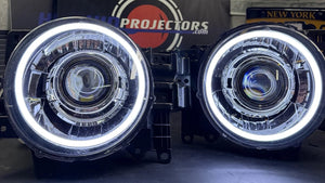 A pair of aftermarket car headlights with bright white LED halo rings, displayed on a dark surface in front of a branded black sign that reads 'HEADLIGHTS BY PROJECTORS'.